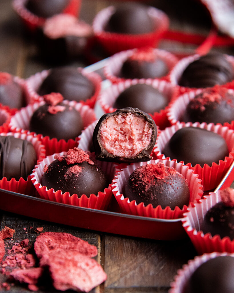 A heart shaped tin full of strawberry cream chocolates. One has a bite out of it showing the strawberry center.