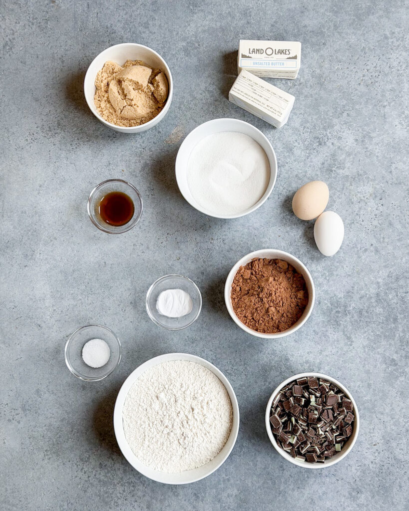 Small bowls of the ingredients needed to make Chocolate Mint Cookies, like flour, cocoa powder, andes mints, sugar, and butter. 