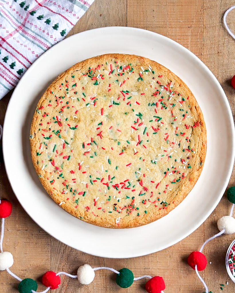 A Christmas cookie cake with Christmas sprinkles on a white plate. 