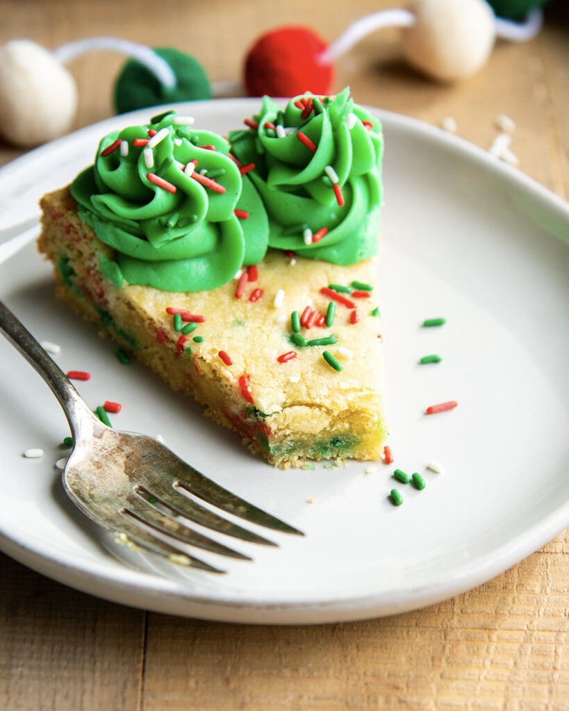 A slice of a Christmas Sugar Cookie Cake on a plate with green buttercream on the edges and red, white, and green sprinkles, with one bite cut out of it.