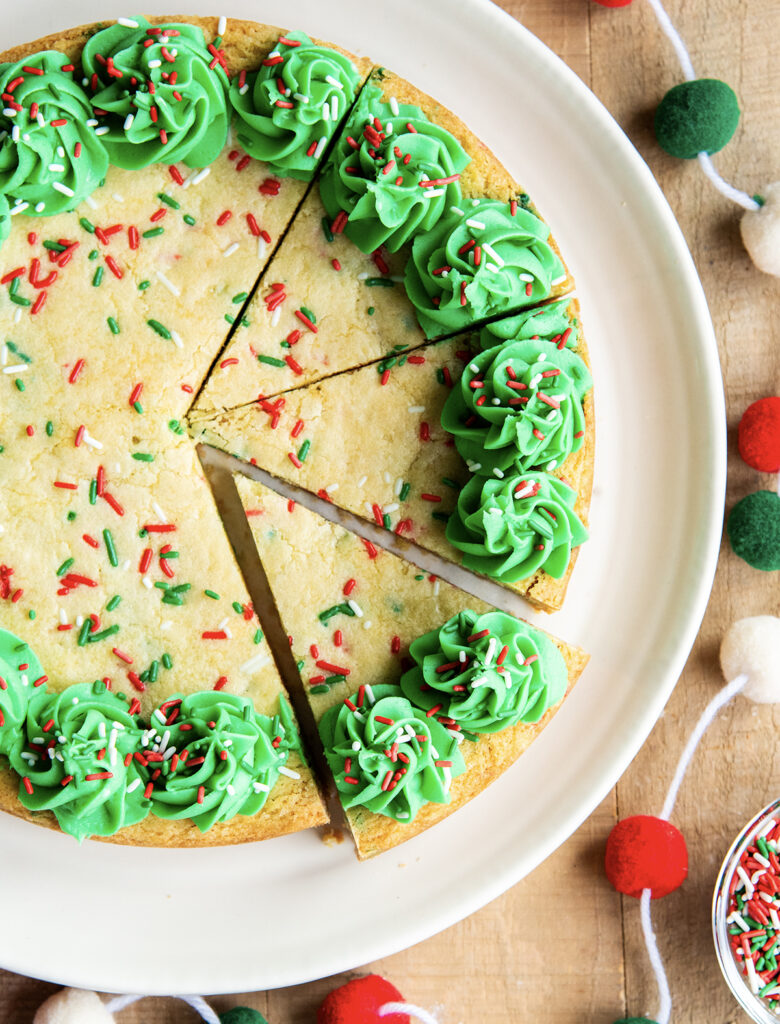 An above view of a Christmas Sugar Cookie Cake on a plate with Christmas sprinkles and green buttercream frosting on the sides. The cake has three slices cut out of it.