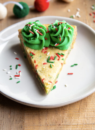 A slice of a Christmas Sugar Cookie Cake on a plate with green buttercream on the edges and red, white, and green sprinkles.