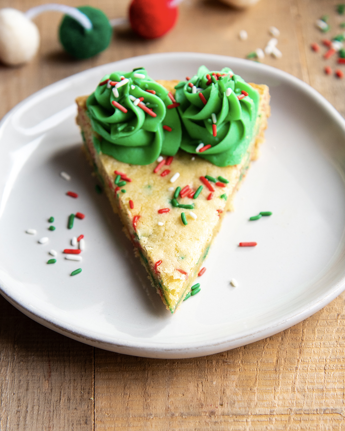 A slice of a Christmas Sugar Cookie Cake on a plate with green buttercream on the edges and red, white, and green sprinkles.