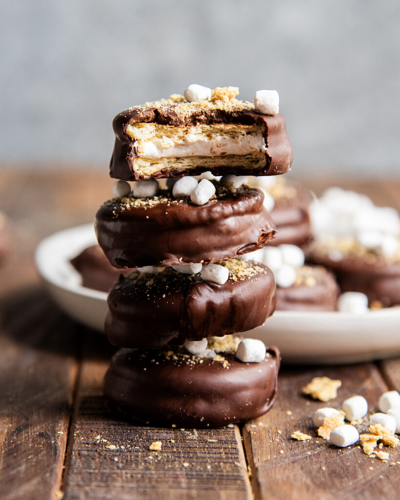 A pile of chocolate covered crackers topped with mini marshmallows and graham cracker crumbs, the top one has a bite out of it showing marshmallow fluff in the middle.