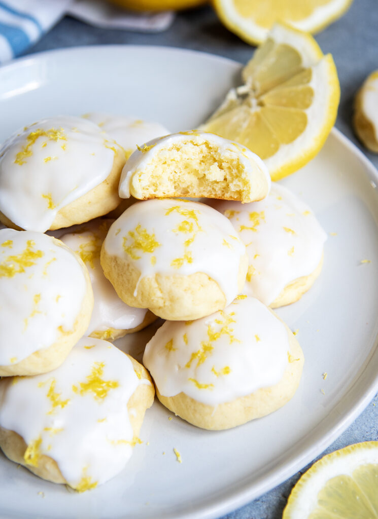 A close up of Italian Lemon Ricotta Cookies topped with white icing and lemon zest on a white plate, one has a bite out of it.