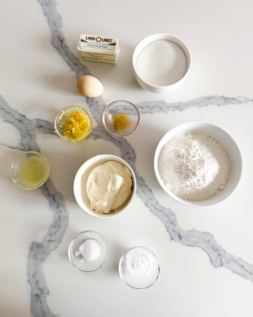 The ingredients needed to make lemon ricotta cookies in small bowls. There is flour, sugar, lemon zest, an egg, butter, and more. 