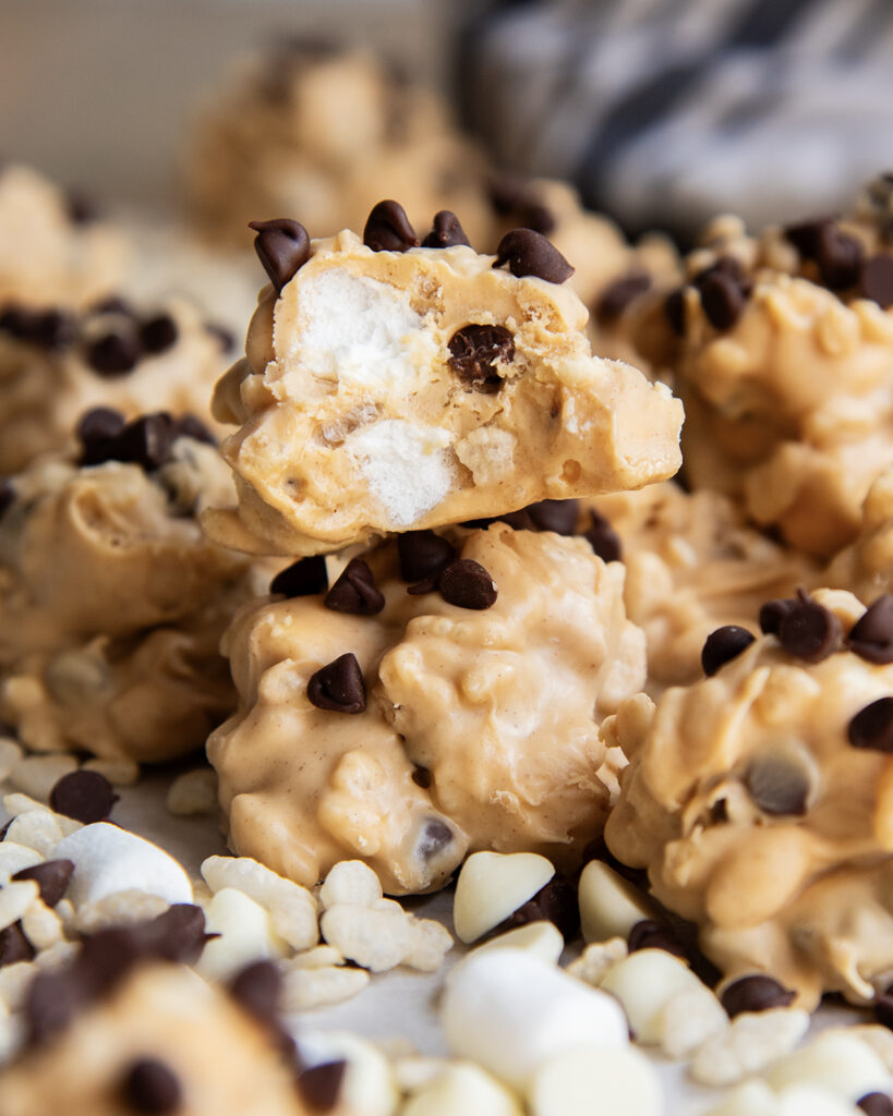 Two peanut butter white chocolate candy clumps, one has a bite out of it with mini marshmallows in it.
