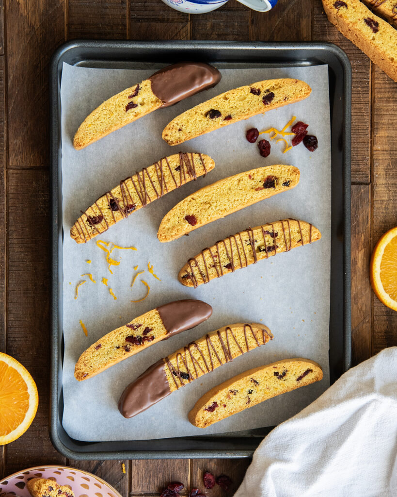 A cookie pan topped with cranberry orange biscotti, some are dipped half way in chocolate, and some are drizzled with chocolate.