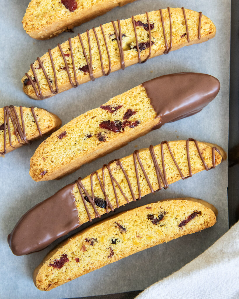 A cookie pan topped with cranberry orange biscotti, some are dipped half way in chocolate, and some are drizzled with chocolate.