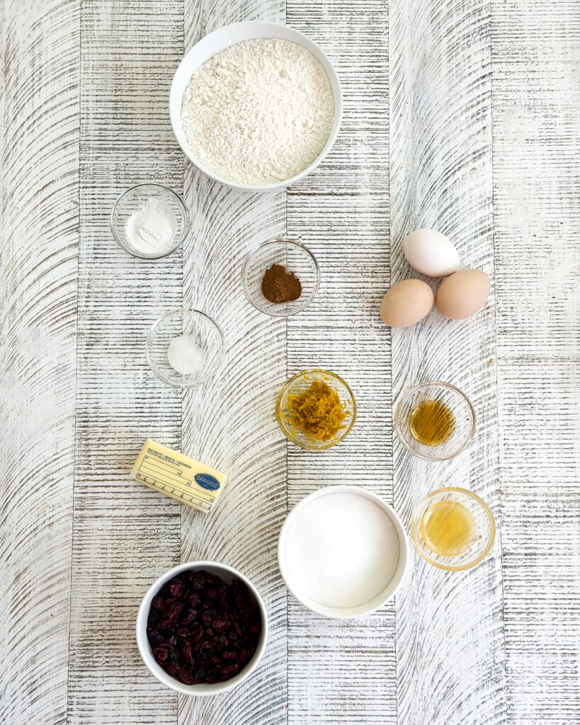 The ingredients needed to make cranberry orange biscotti in small bowls, like butter, flour, baking powder, cinnamon, orange zest and eggs. 