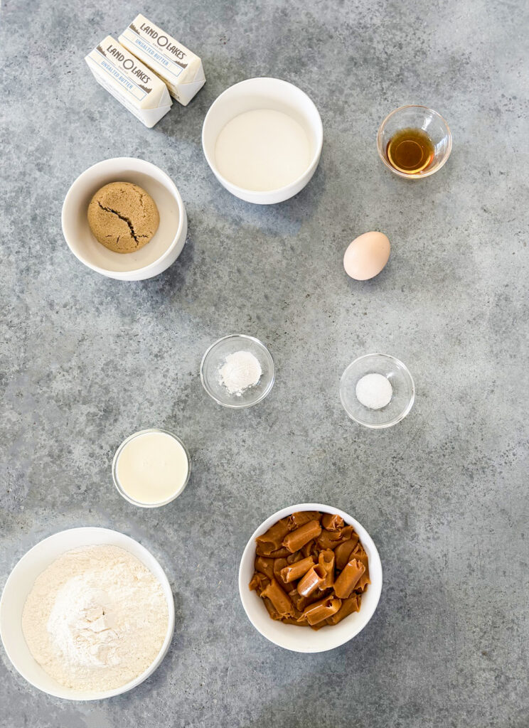 Small bowls of the ingredients needed to make caramel shortbread bars, including sugars, caramels, flour, butter, an egg, and vanilla.