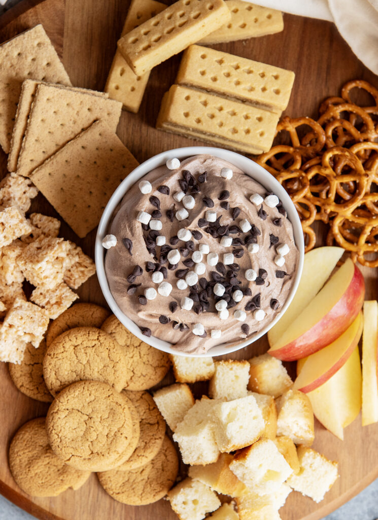 A wooden board topped with cookies and other dippers and a bowl of hot chocolate dip.
