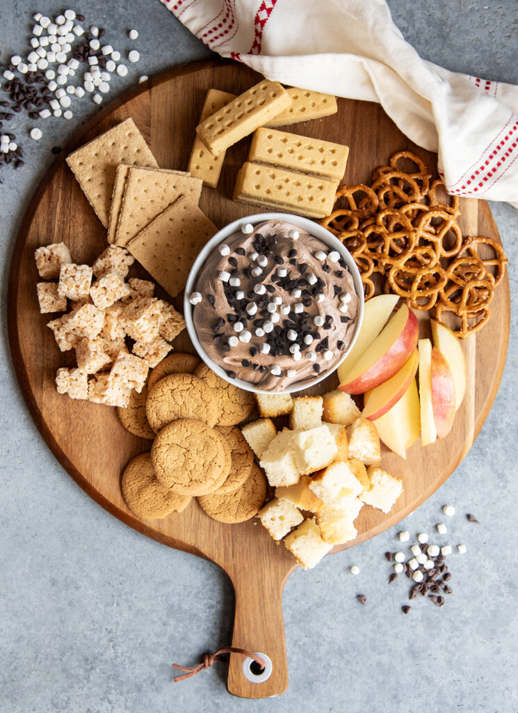 A wooden board topped with cookies and other dippers and a bowl of hot chocolate dip.
