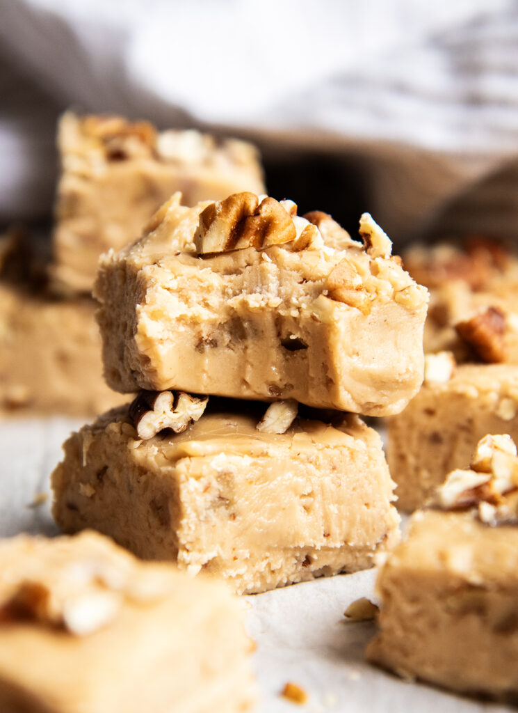 A stack of two pieces of pecan brown sugar penuche fudge topped with chopped pecans, the top piece has a bite out of it.