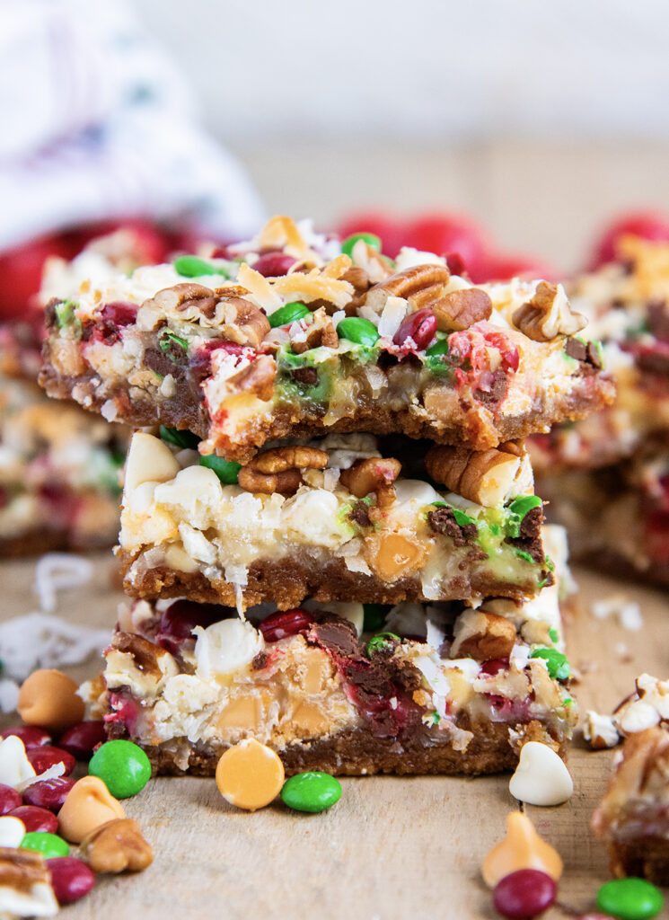 A stack of three Christmas Magic Cookie bars with butterscotch chips, m&ms, and coconut. The top bar has a bite out of it. 