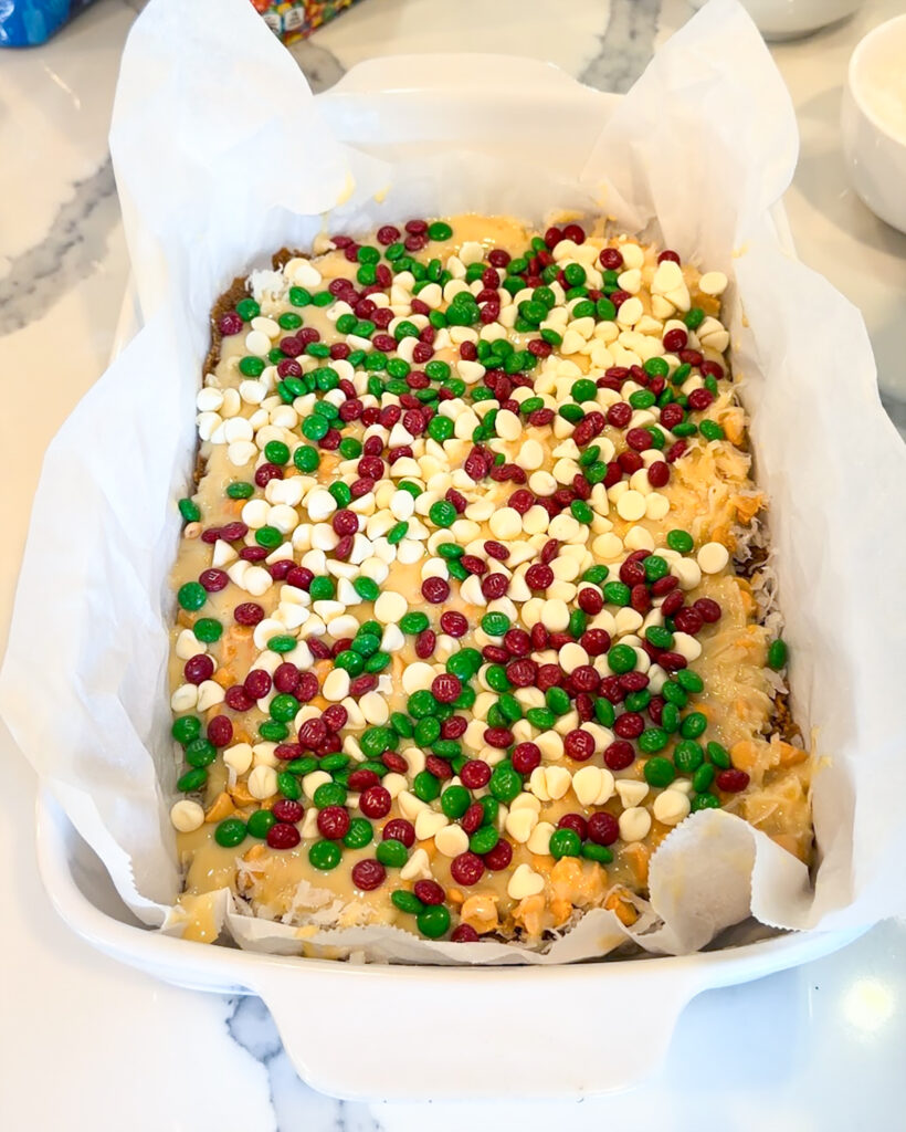 Uncooked magic cookie bars topped with white chocolate chips and m&ms.