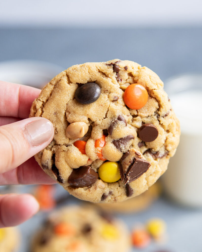 A hand holding a peanut butter cookie stuffed with peanut butter cups and reeses pieces.