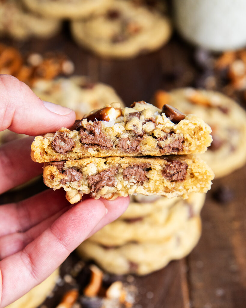 A hand holding two halves of a pretzel chocolate chip cookie showing the chocolate chips in the middle.