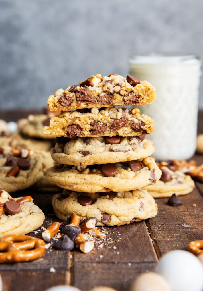 A stack of four drop style cookies filled with chocolate chips and pretzel pieces, the top cookie is broken in half showing the inside of the cookies.
